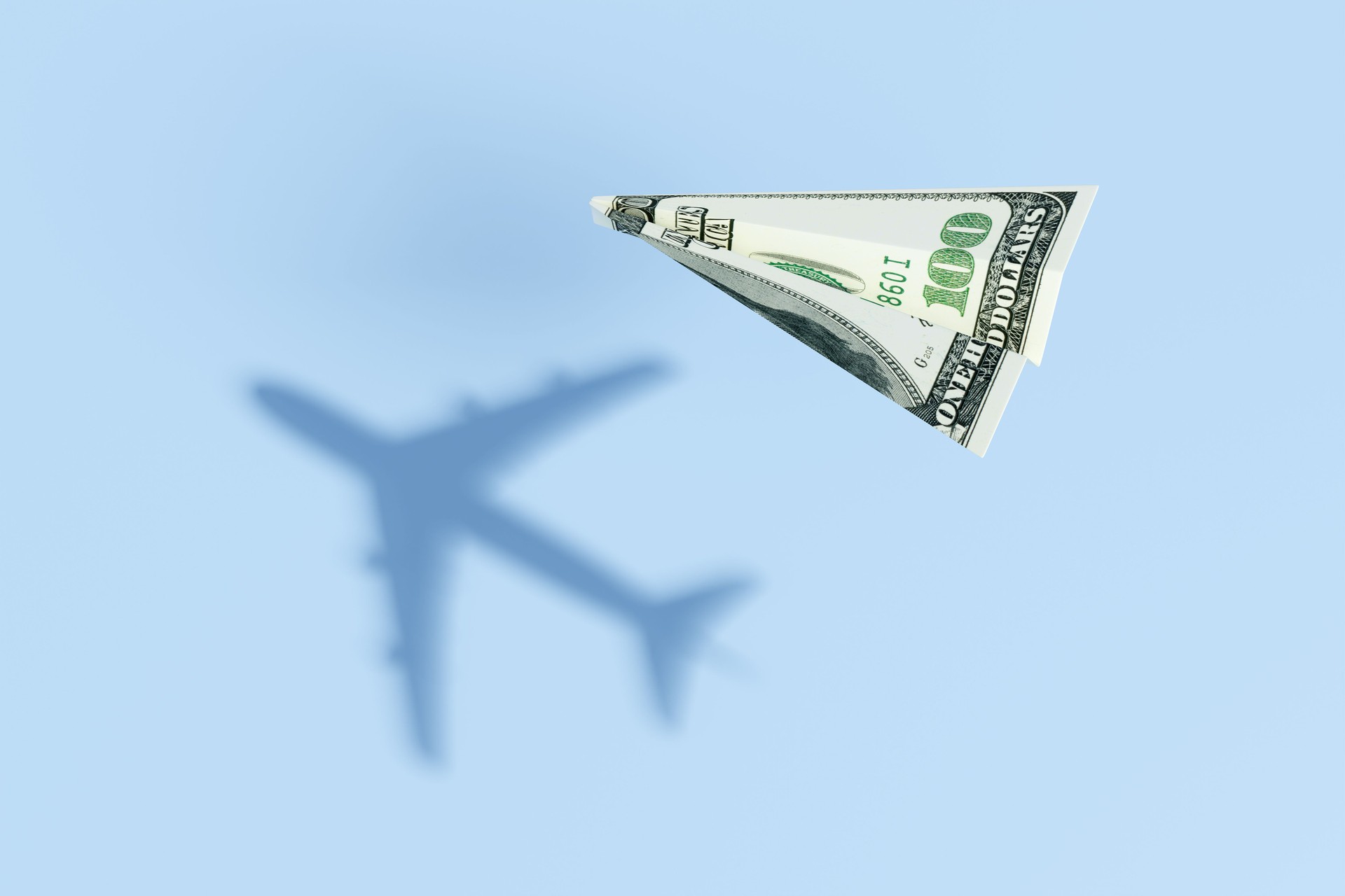 Avión de papel hecho de un billete de 100 dólares estadounidenses que vuela sobre un fondo marino azul y proyecta una sombra de vuelo comercial realista. Ilustración del concepto de presupuesto de viaje y precios de boletos aéreos